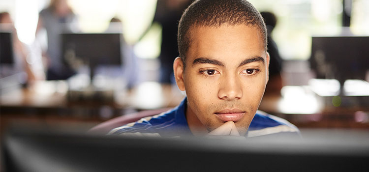 man pensively works on desktop computer in classroom with blurred computers and students behind him